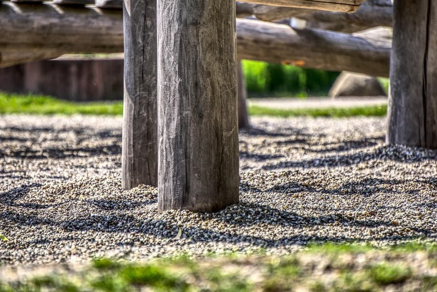 Nahaufnahme von dicken Holzpfosten eines Spielplatzgeräts, die in einem Untergrund aus feinem Kies stehen, mit unscharfem Hintergrund aus Rasen und weiteren Holzelementen.